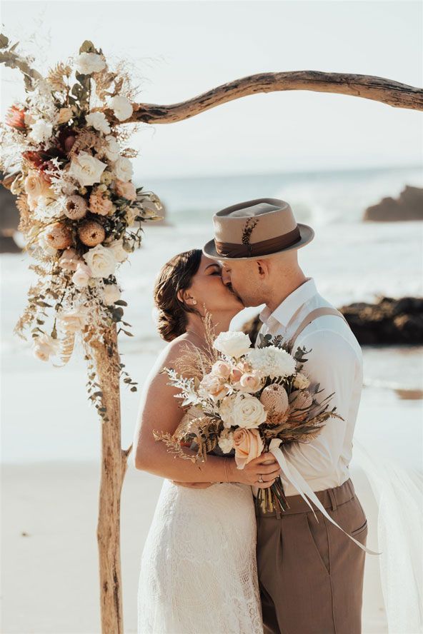 A bride and groom kissing under a wooden arch on the beach.