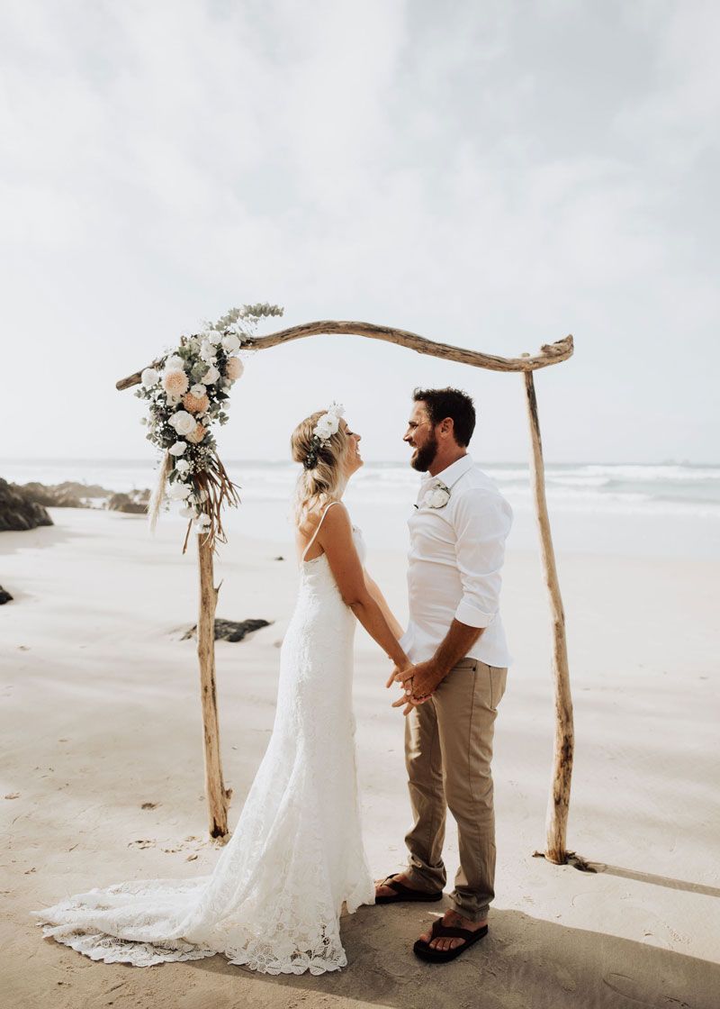 A bride and groom are holding hands on the beach under a wooden arch.