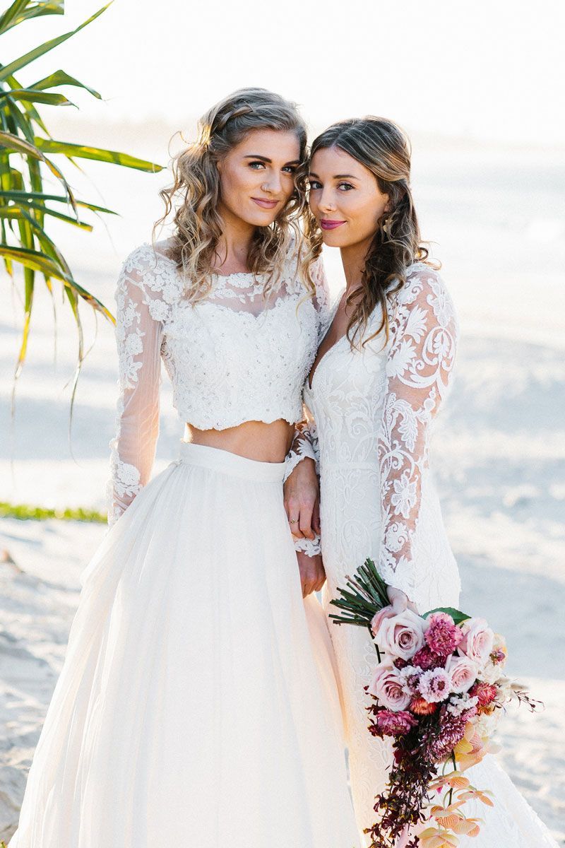 Two women in white dresses are posing for a picture on the beach.