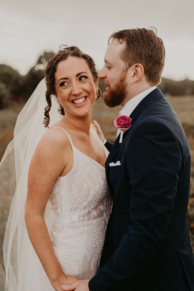 A bride and groom are standing next to each other in a field and smiling at each other.