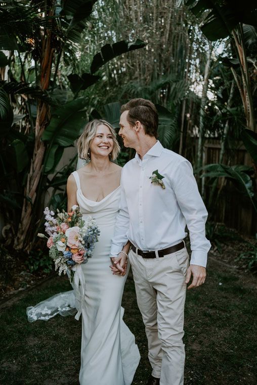A bride and groom are holding hands and smiling while walking in a garden.