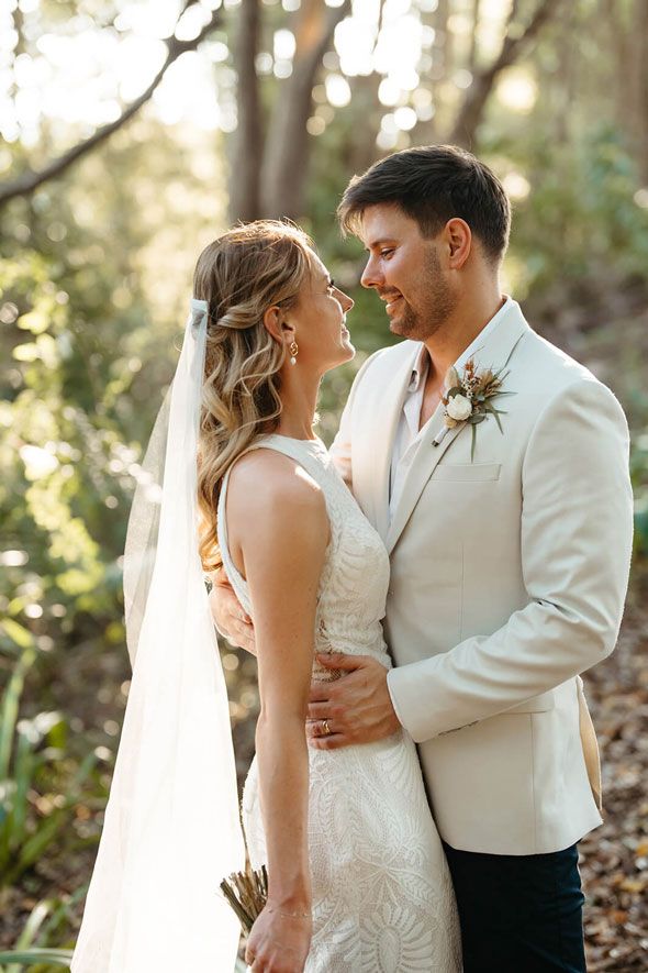 A bride and groom are standing next to each other in the woods and looking at each other.