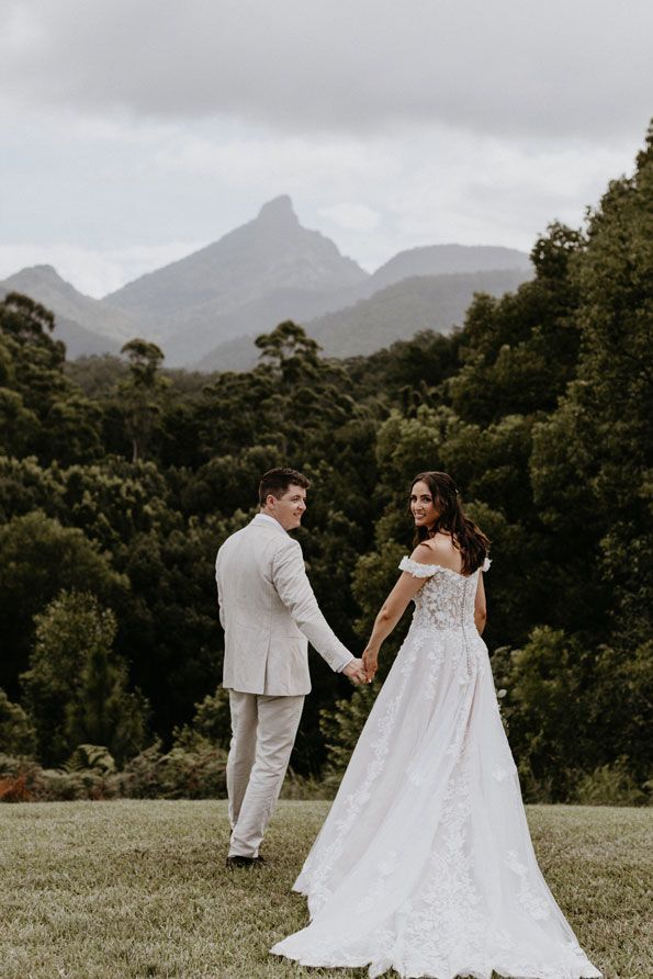 A bride and groom are holding hands in a field with mountains in the background.