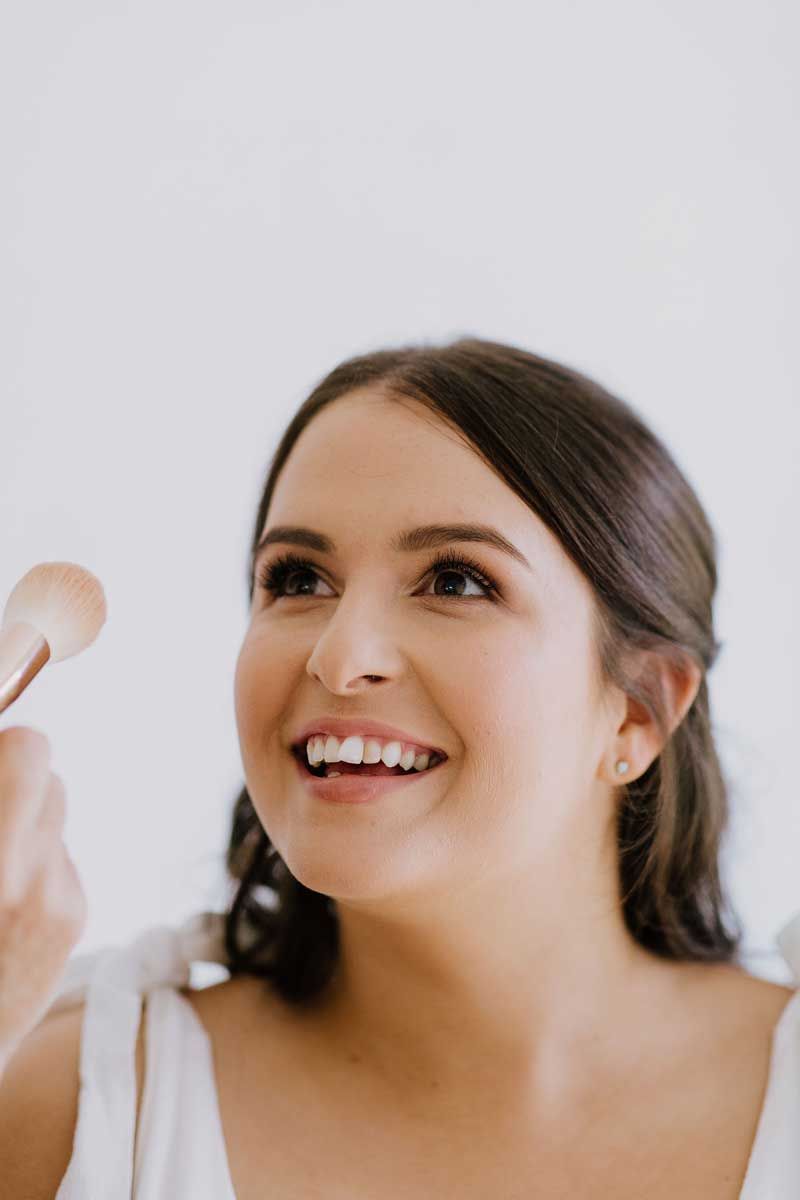 A woman is smiling while applying makeup in front of a mirror.