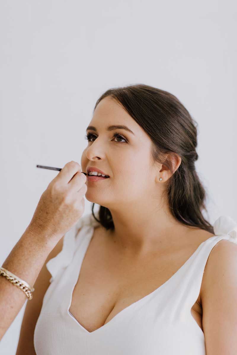 A woman in a white dress is getting her makeup done by a makeup artist.