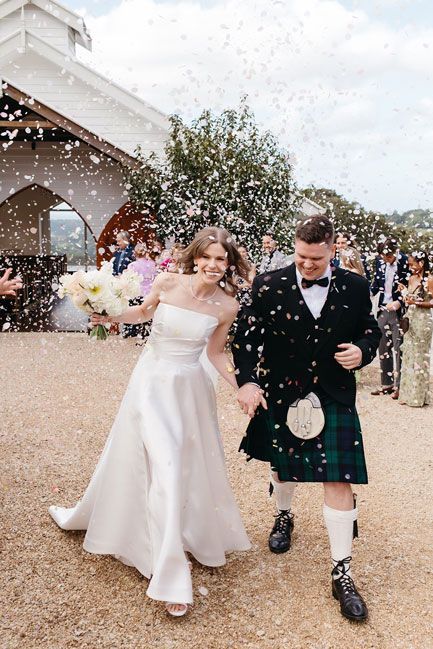 A bride and groom in a kilt are walking away from a church covered in confetti.