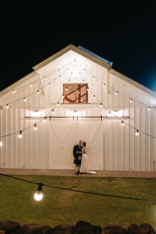 A bride and groom are standing in front of a white barn at night.