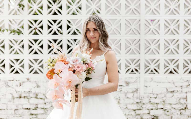A bride in a white dress is holding a bouquet of flowers in front of a white brick wall.