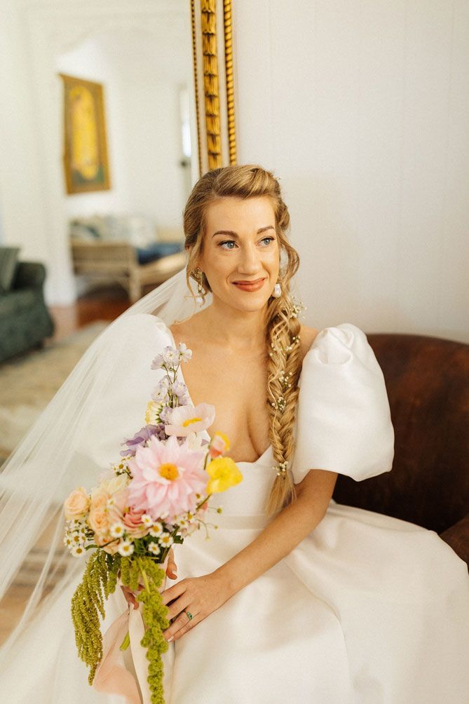 A bride in a wedding dress is sitting on a couch holding a bouquet of flowers.