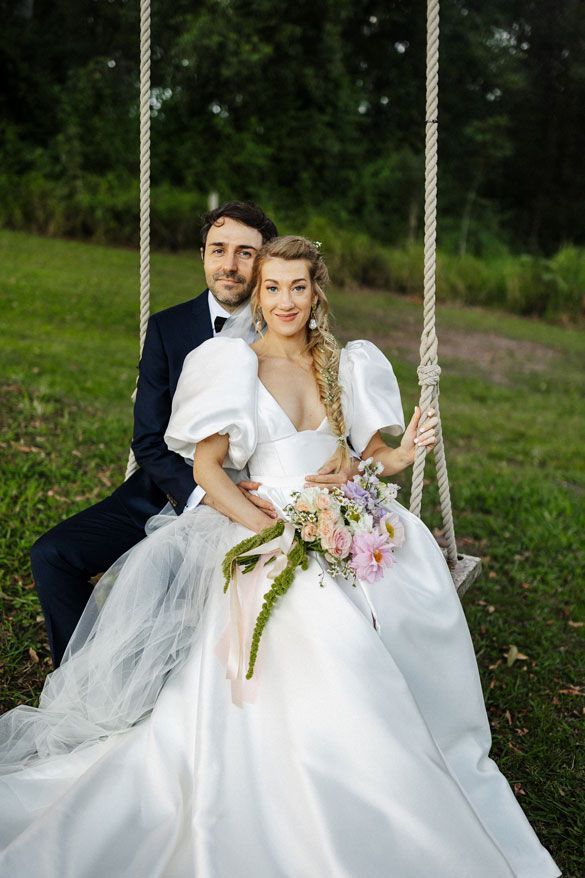 A bride and groom are sitting on a swing in the grass.