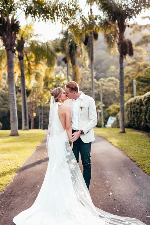 A bride and groom are kissing on a path surrounded by palm trees.