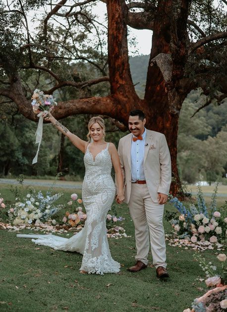 A bride and groom are standing in front of a tree holding hands.