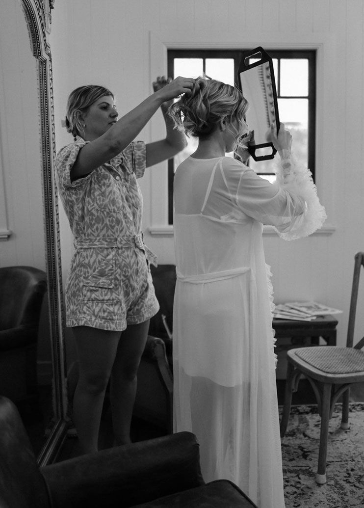 A woman is helping another woman with her hair in front of a mirror.
