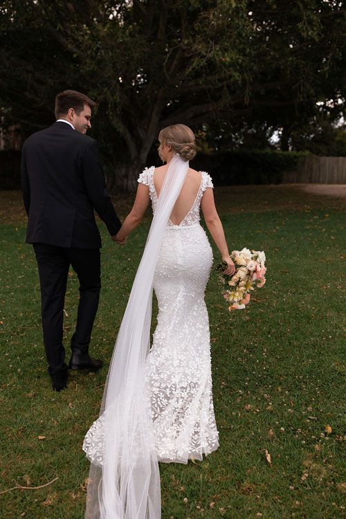 A bride and groom are walking through a grassy field holding hands.