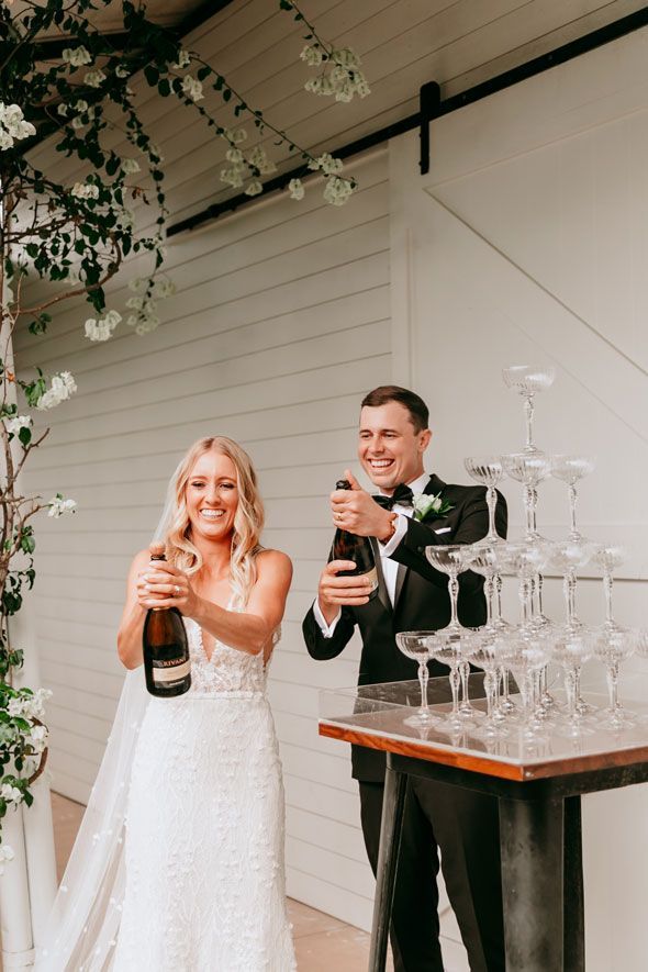 A bride and groom are toasting with champagne in front of a pyramid of champagne glasses.