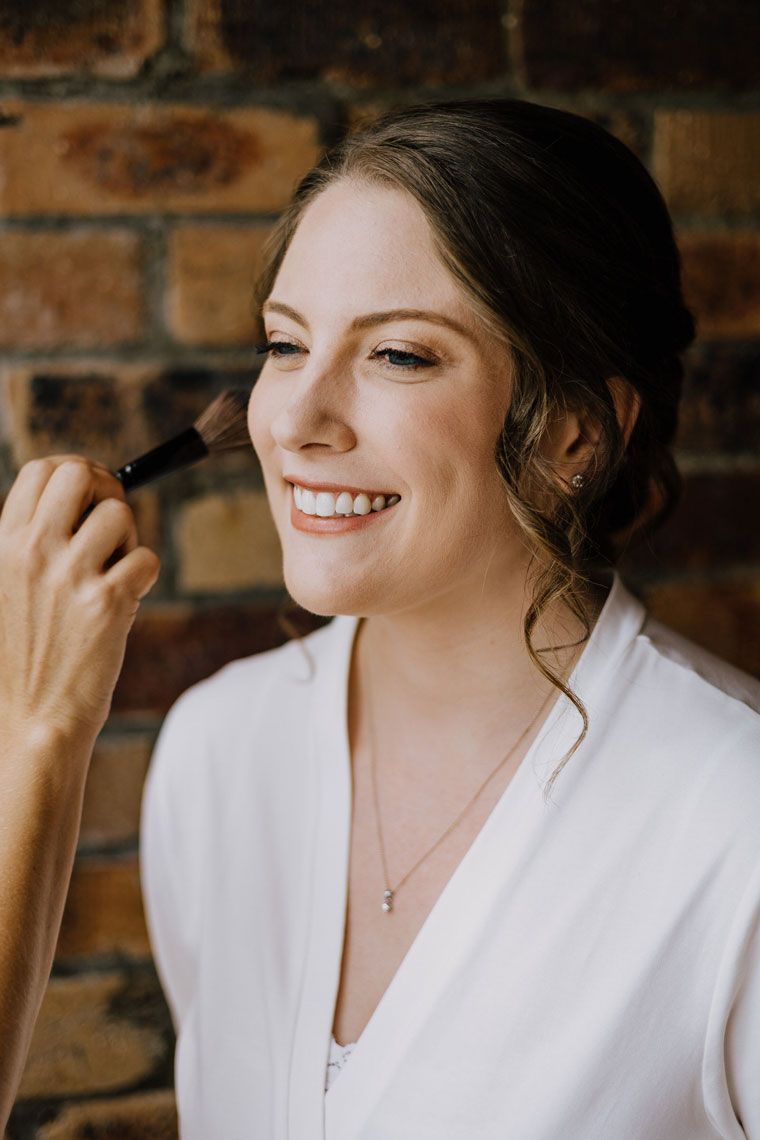 A woman is smiling while getting her makeup done in front of a brick wall.