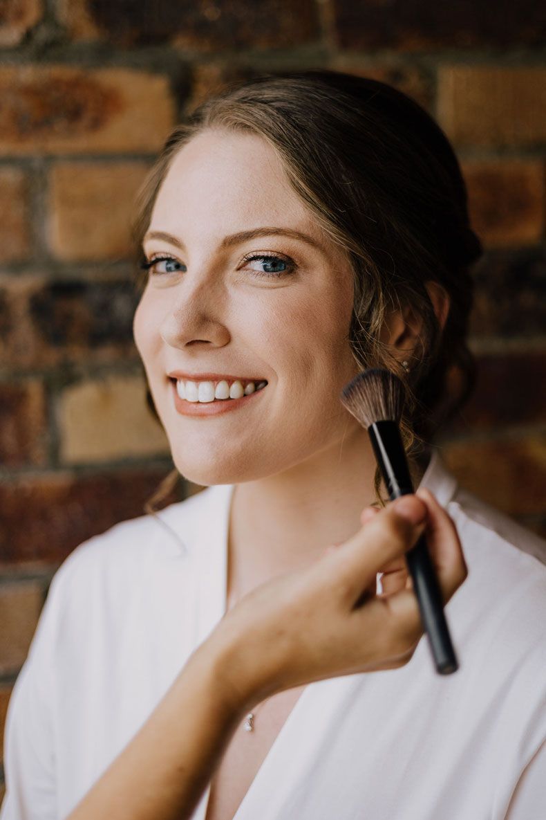 A woman is getting her makeup done by a makeup artist in front of a brick wall.
