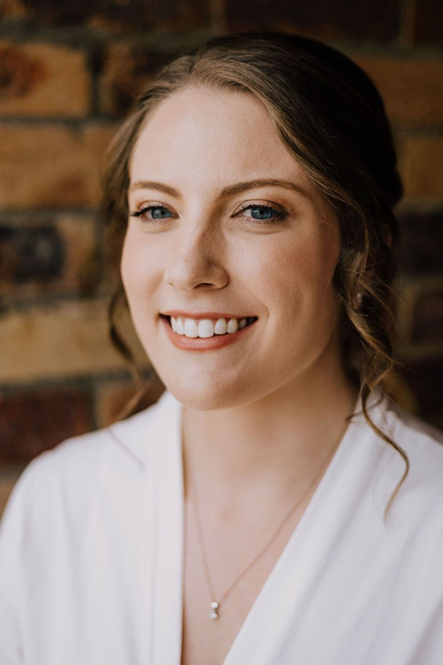 A woman in a white robe is smiling in front of a brick wall.