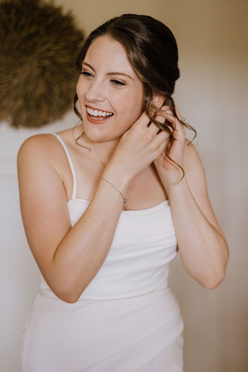 A woman in a white dress is putting on her earrings and smiling.