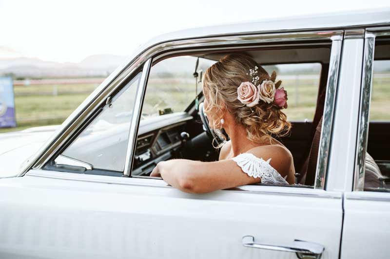 A bride is sitting in a white car looking out the window.