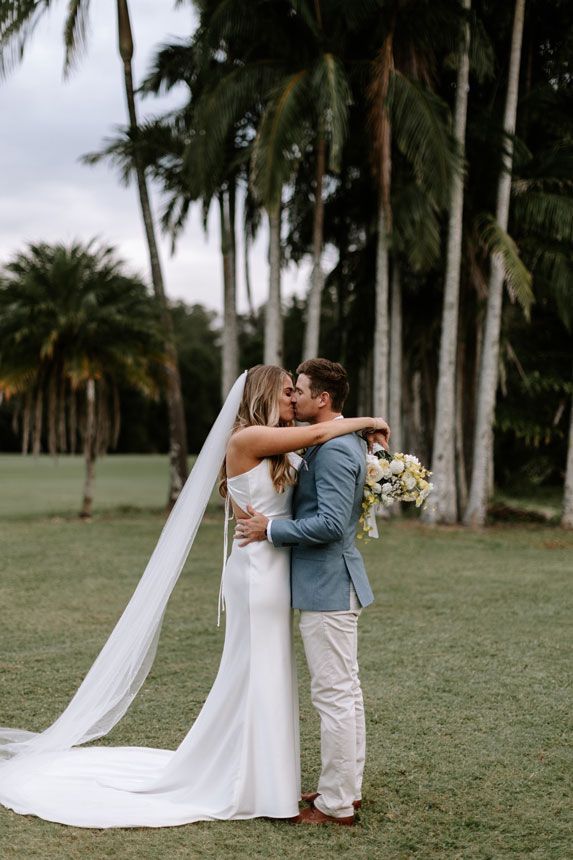 A bride and groom are kissing in a field with palm trees in the background.