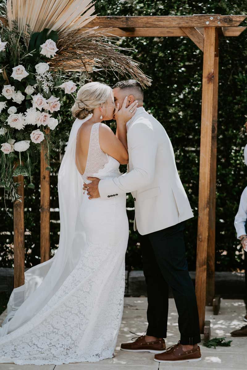 A bride and groom are kissing at their wedding ceremony under a wooden arch.