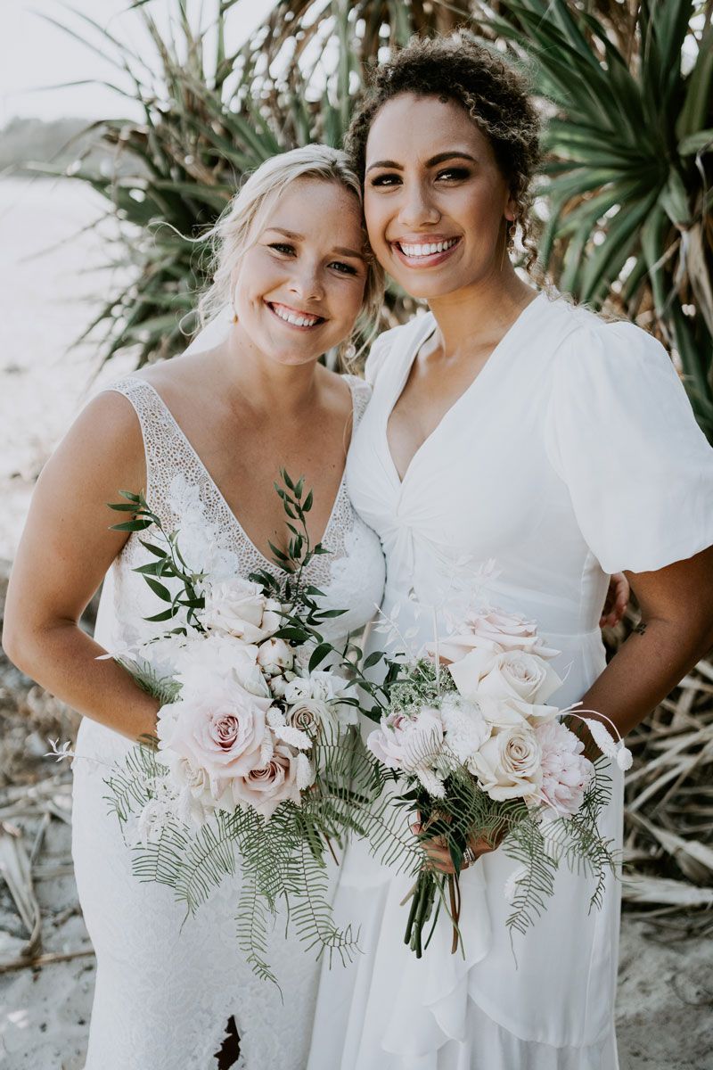 Two women in white dresses are posing for a picture while holding bouquets of flowers.
