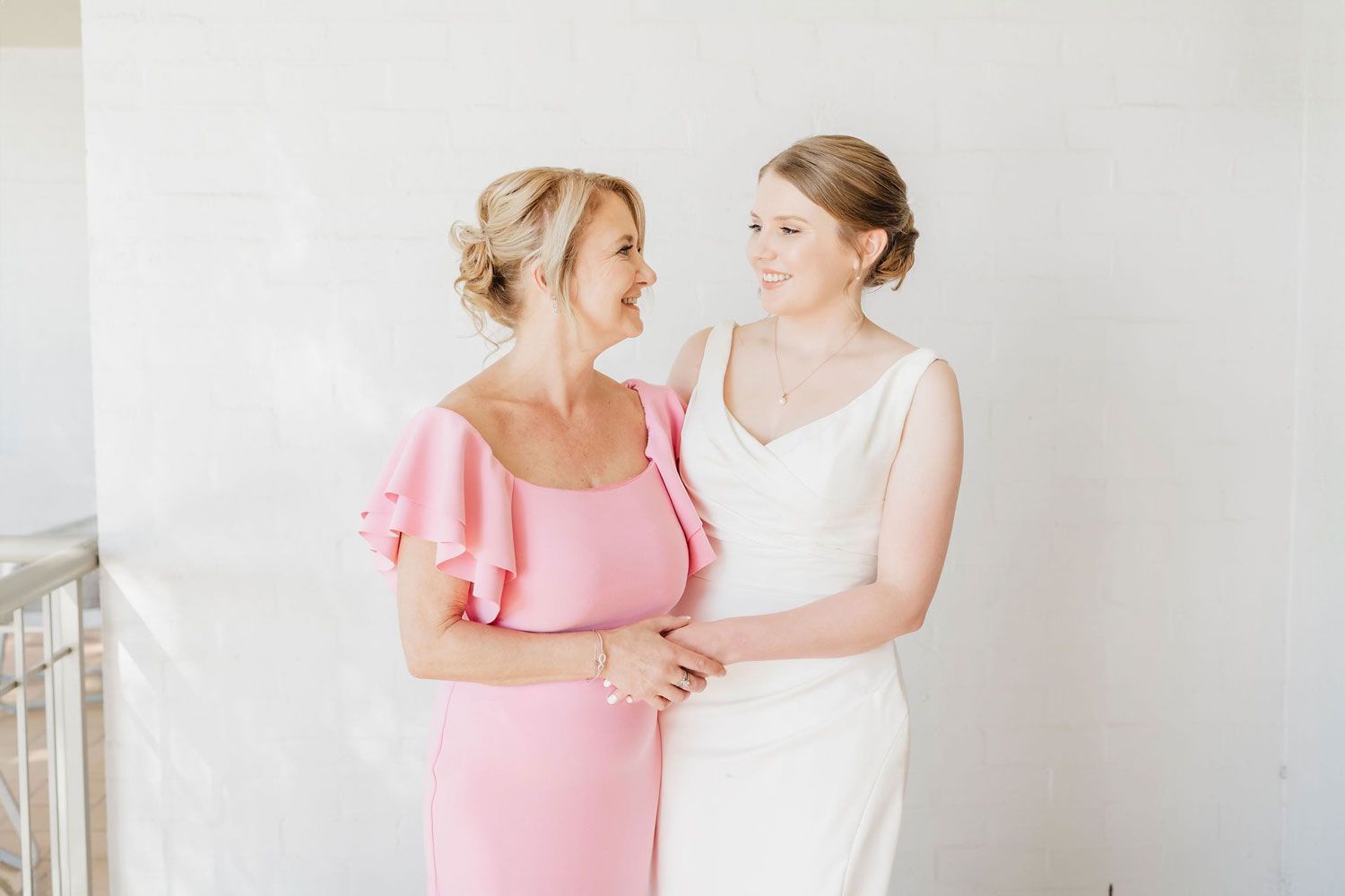 A bride and her mother are standing next to each other in front of a white wall.