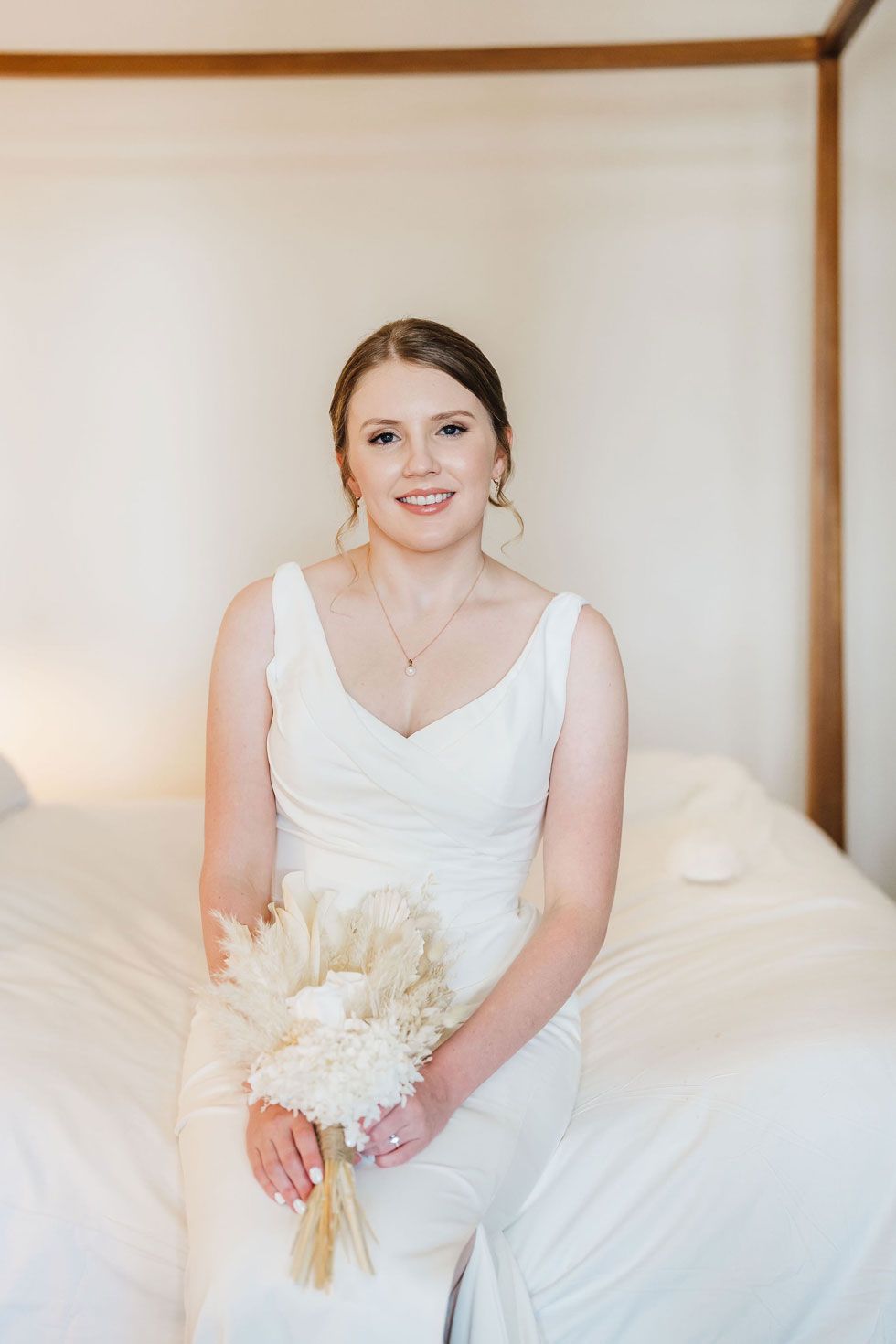 A bride in a white dress is sitting on a bed holding a bouquet of flowers.