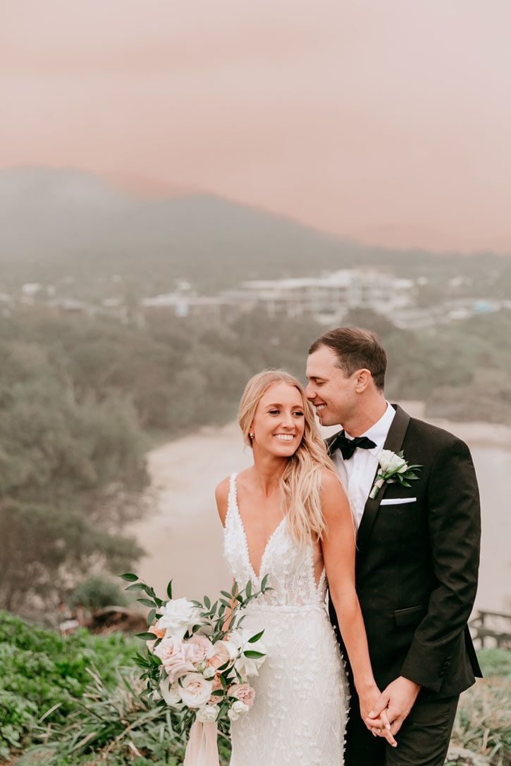 A bride and groom are standing next to each other on top of a hill holding hands.