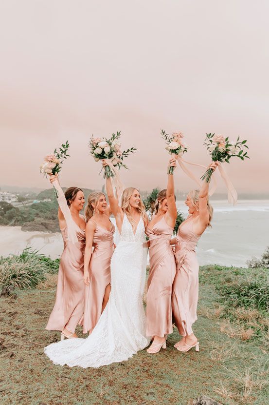A bride and her bridesmaids are standing on top of a hill holding their bouquets in the air.