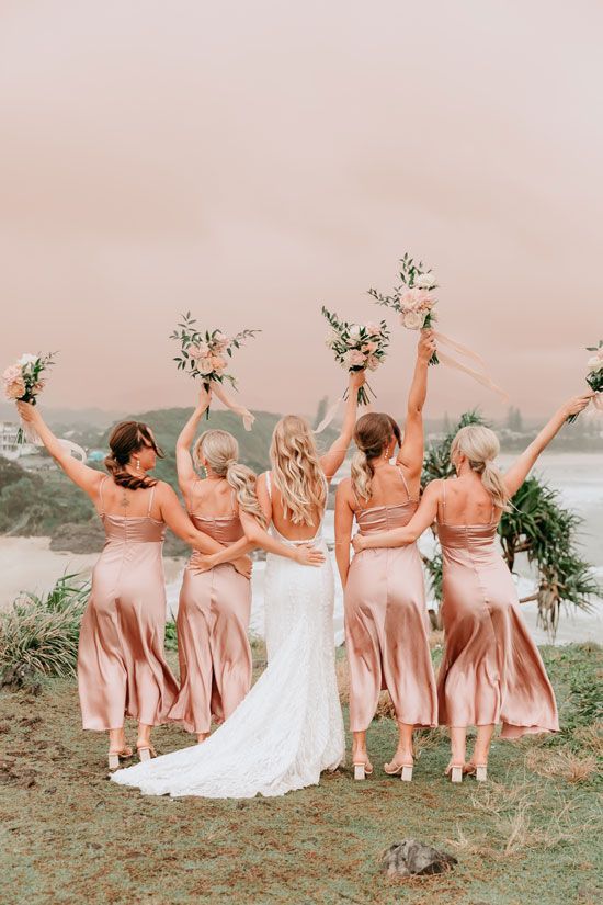 A bride and her bridesmaids are standing on top of a hill holding flowers in the air.