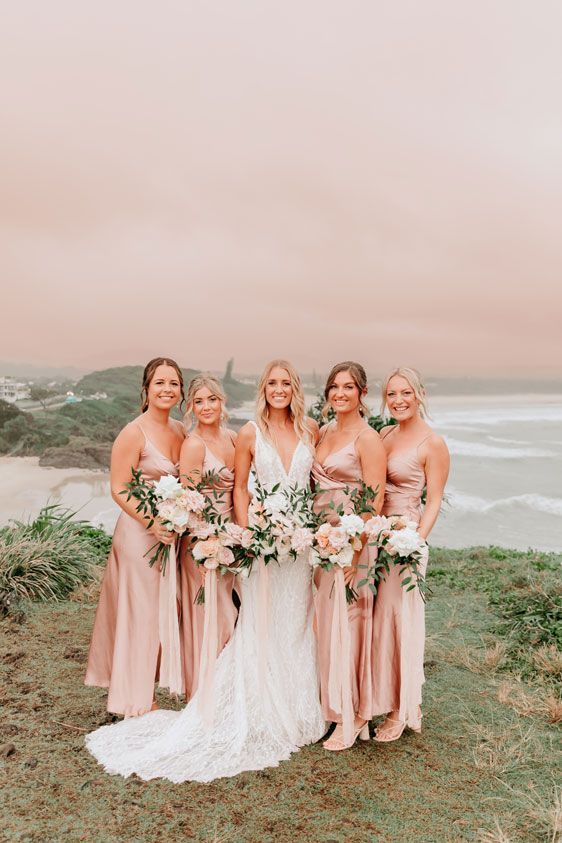 A bride and her bridesmaids are posing for a picture on a hill overlooking the ocean.