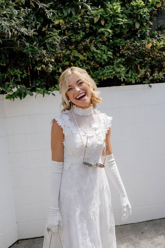 A woman in a white dress and white gloves is standing in front of a white wall.