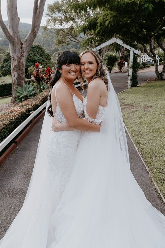 Two women in wedding dresses are hugging each other on a sidewalk.