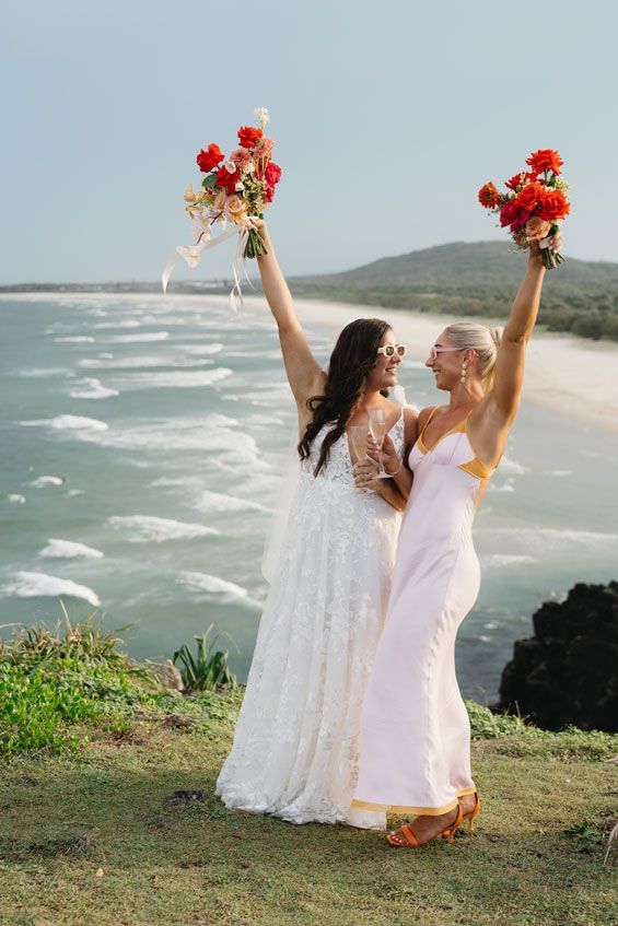 Two brides are standing next to each other on a beach holding their bouquets in the air.