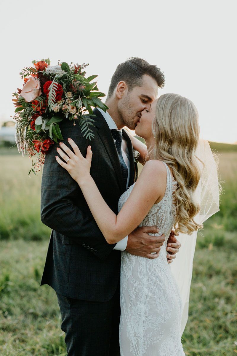 A bride and groom are kissing in a field while the bride is holding a bouquet of flowers.