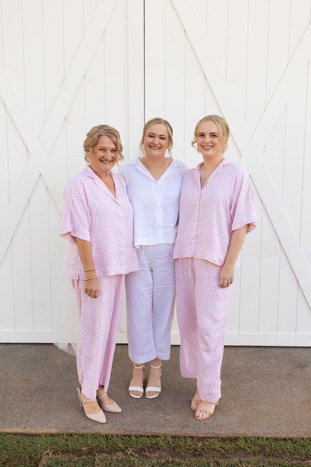Three women in pink pajamas are standing next to each other in front of a white barn.