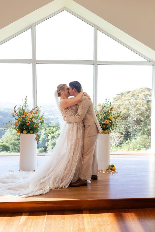 A bride and groom are kissing at their wedding ceremony in front of a large window.