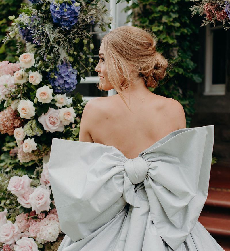 A woman in a wedding dress with a large bow on the back is standing in front of flowers.