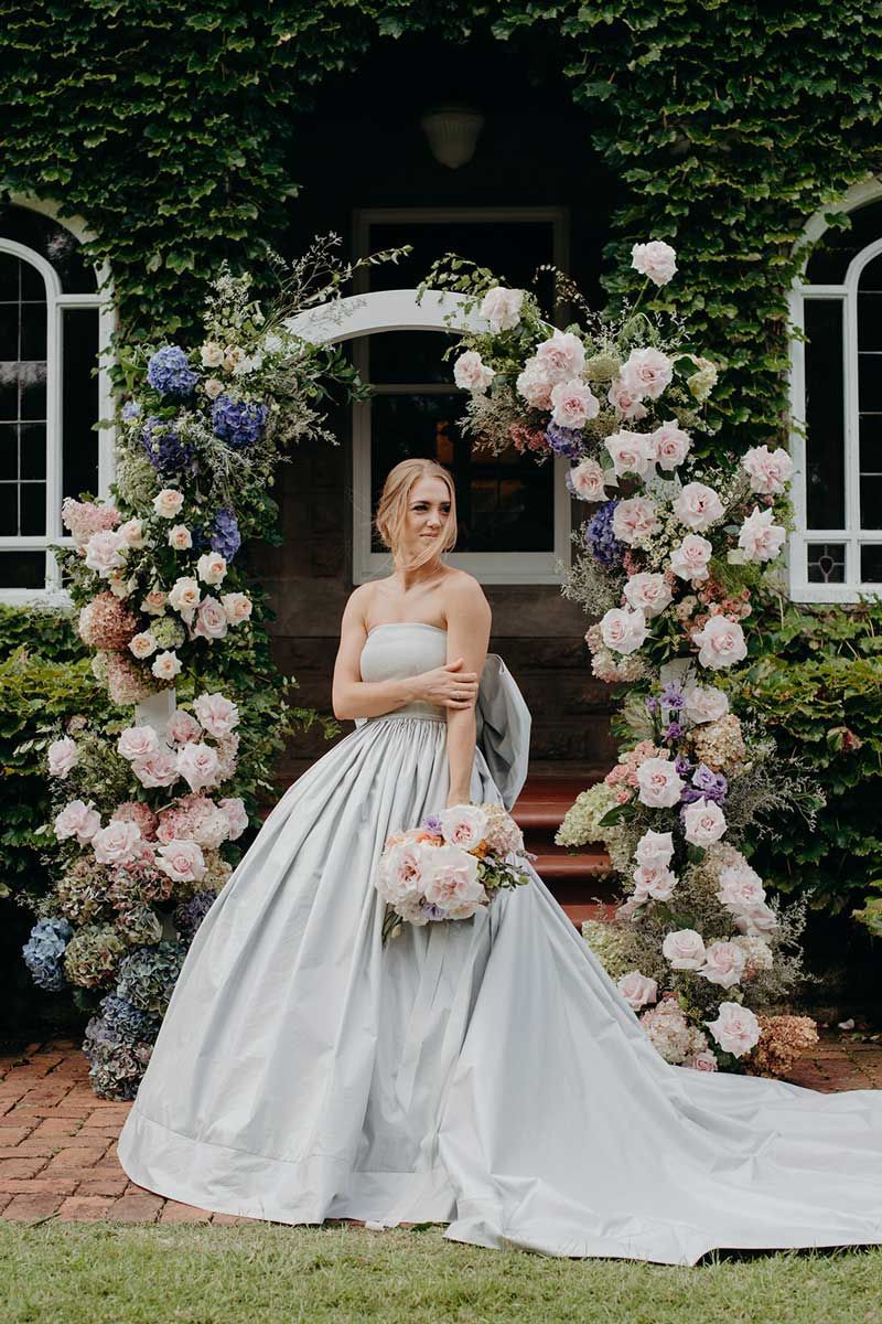 A bride in a light blue wedding dress is standing in front of a floral arch.