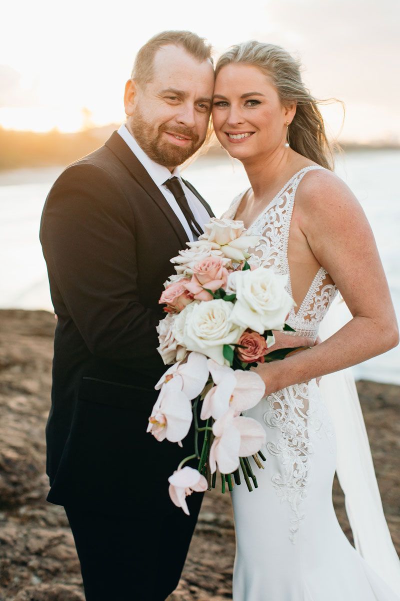 A bride and groom are posing for a picture on the beach . the bride is holding a bouquet of flowers.