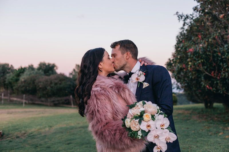 A bride and groom are kissing in a field . the bride is wearing a pink fur coat.