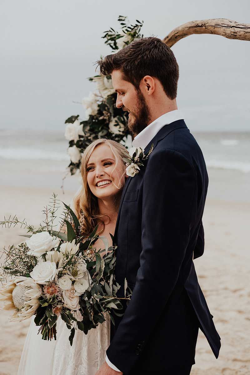 A bride and groom are posing for a picture on the beach.