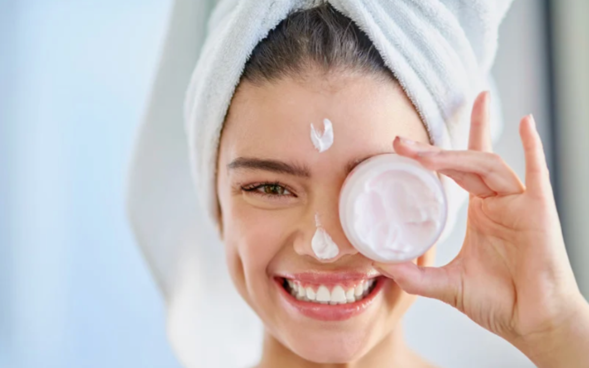 A woman with a towel wrapped around her head is holding a jar of cream in front of her eye.