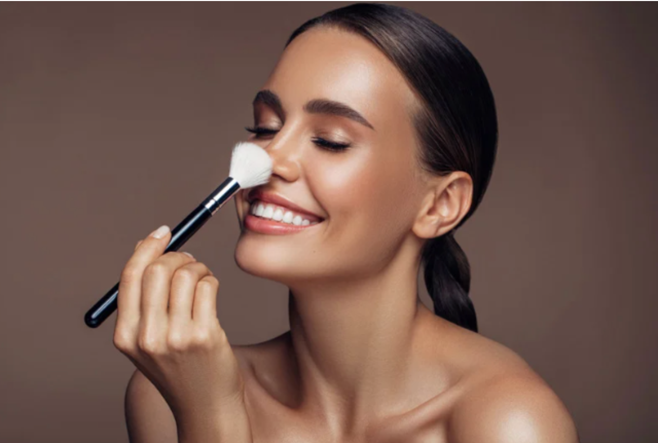 A woman is smiling while applying makeup to her face with a brush.