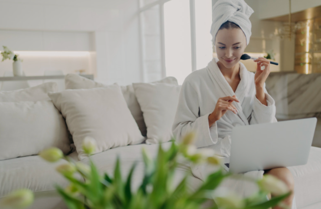 A woman in a bathrobe is sitting on a couch using a laptop and applying makeup.