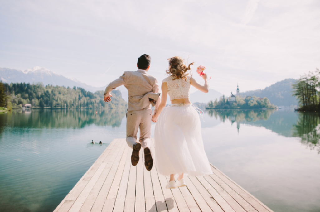 A bride and groom are jumping off a dock into a lake.