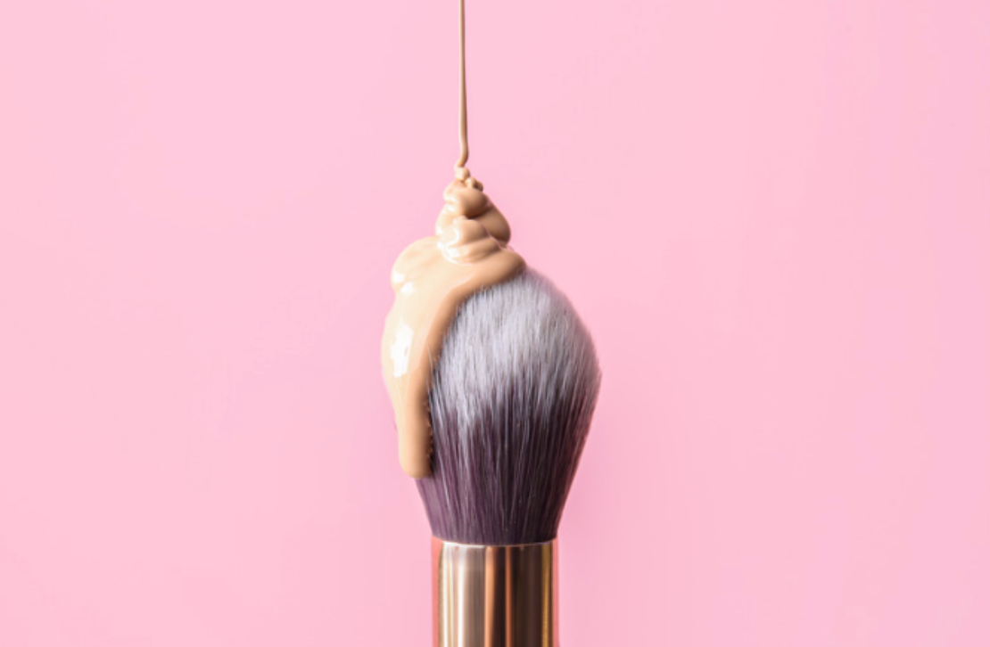 A makeup brush with foundation dripping from it on a pink background.