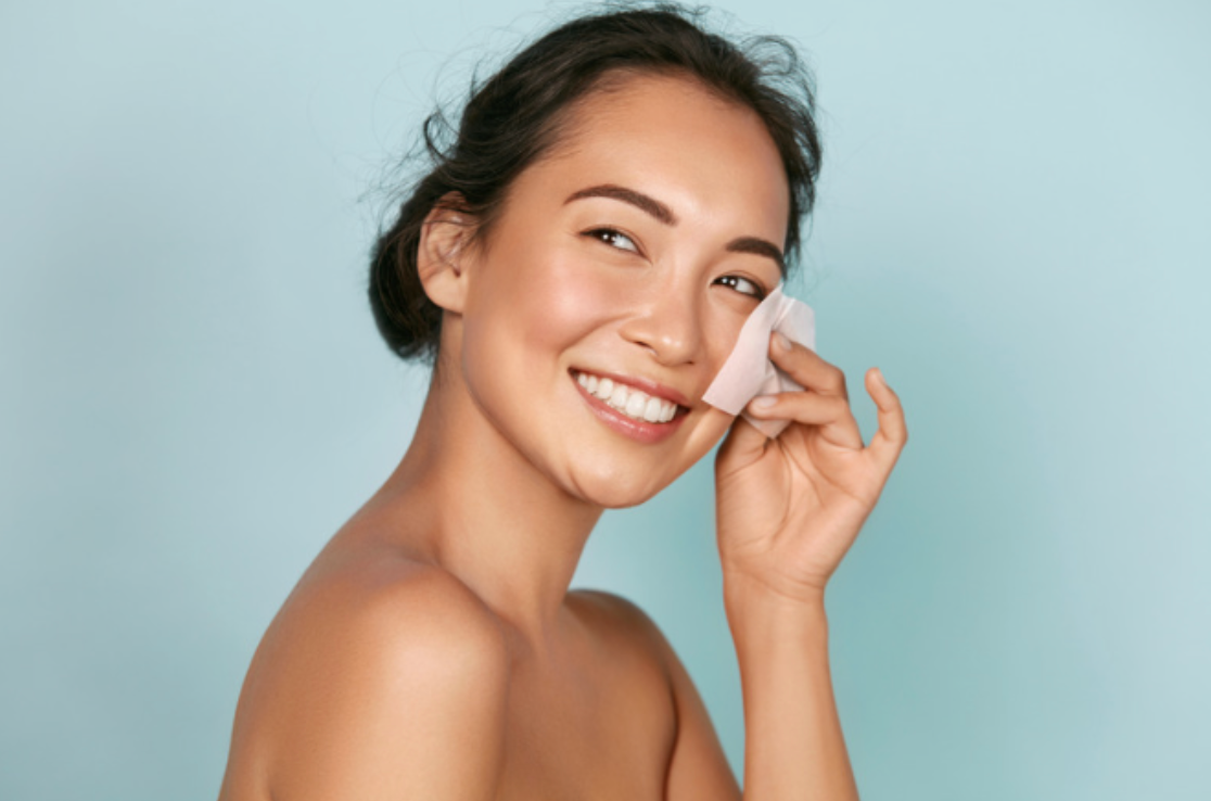 A woman is smiling while cleaning her face with a cotton pad.